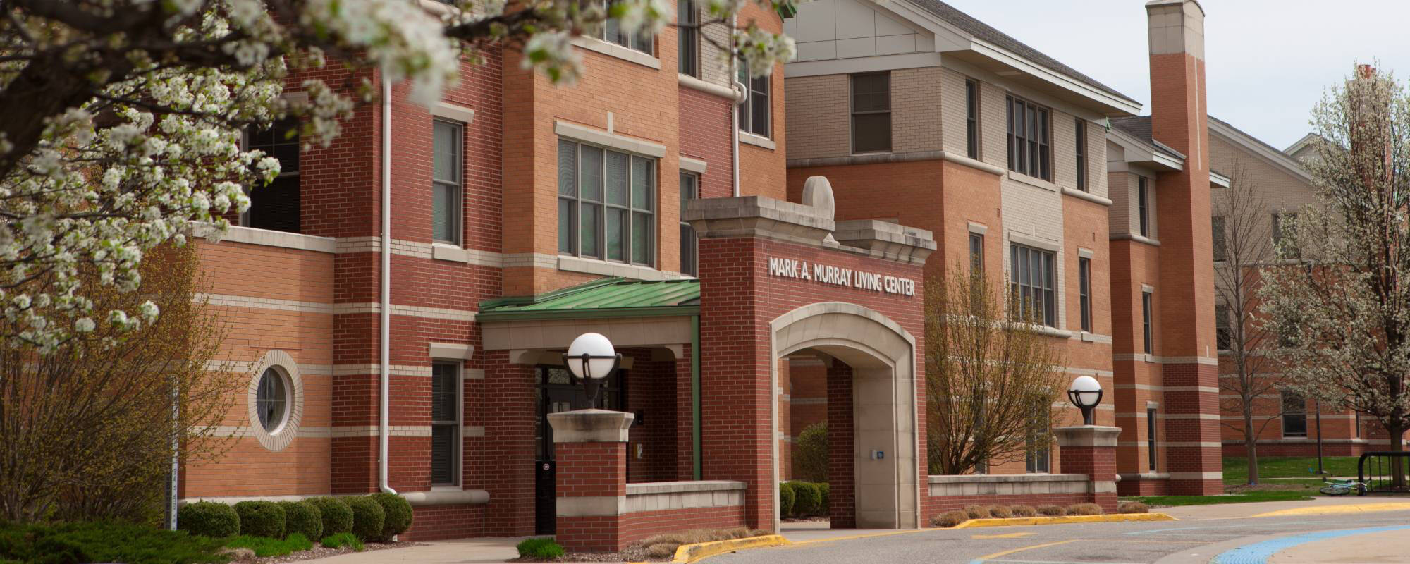 Red-brick building with arched entrance labeled "Mary A. Murray Living Center," flanked by blossoming trees on a bright, clear day.